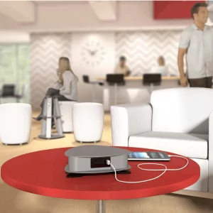 Stand-alone charging station on a red table in a modern office lounge, with people seated in white chairs, providing a solution for powering devices in communal spaces.