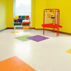 Colorful vinyl composite tile flooring in a bright yellow room, featuring orange, green, purple, and blue patterned tiles, with a red activity table and storage bins in the background, suitable for educational and commercial settings.
