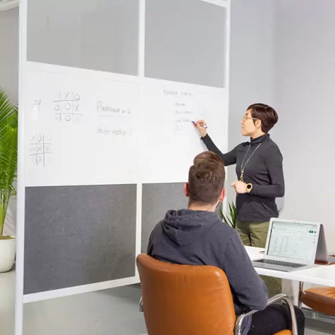 Room divider in a modern office setting with a woman writing on a whiteboard and a man seated at a desk with a laptop, illustrating a collaborative workspace environment.
