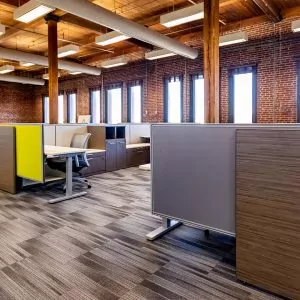 Modern office cubicles in a spacious workspace featuring wooden beams, large windows, and a mix of gray and yellow partitions, emphasizing a collaborative environment.